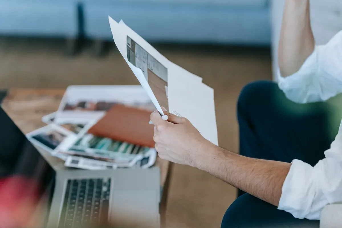 A person reviewing financial documents and a laptop at a home desk near a window