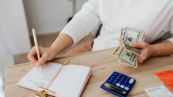 Person writing in a budget notebook while holding cash and a calculator, illustrating moneymaxxing habits