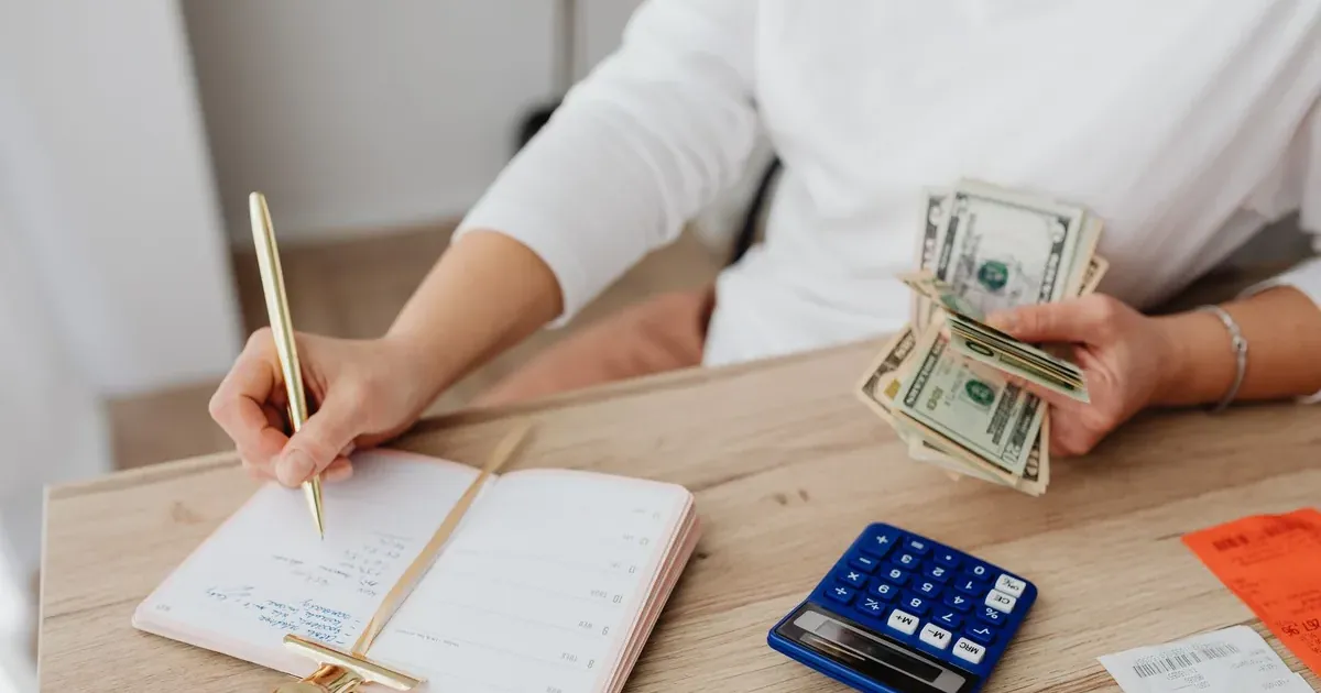 Person writing in a budget notebook while holding cash and a calculator, illustrating moneymaxxing habits