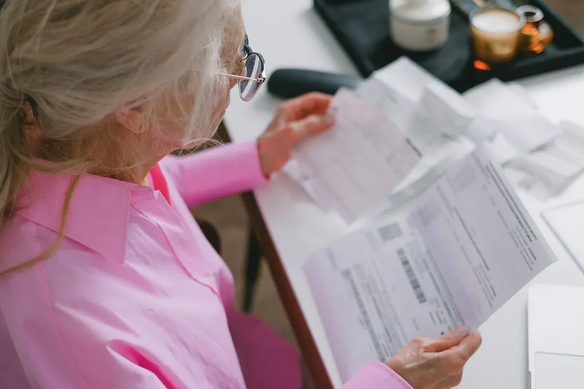 Elderly woman reviewing bills and documents at a kitchen table, representing tracking income against rising costs
